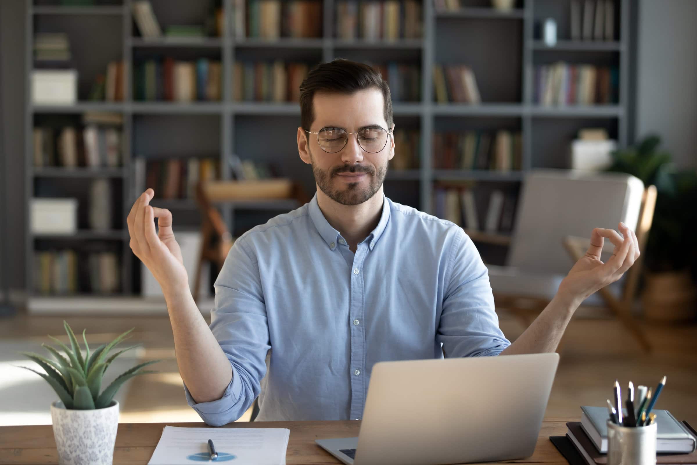 Calm young Caucasian male employee sit at home office workplace practice yoga relieve negative emotions. Relaxed mindful man meditate with mudra hands with eyes closed. Stress free, balance concept.