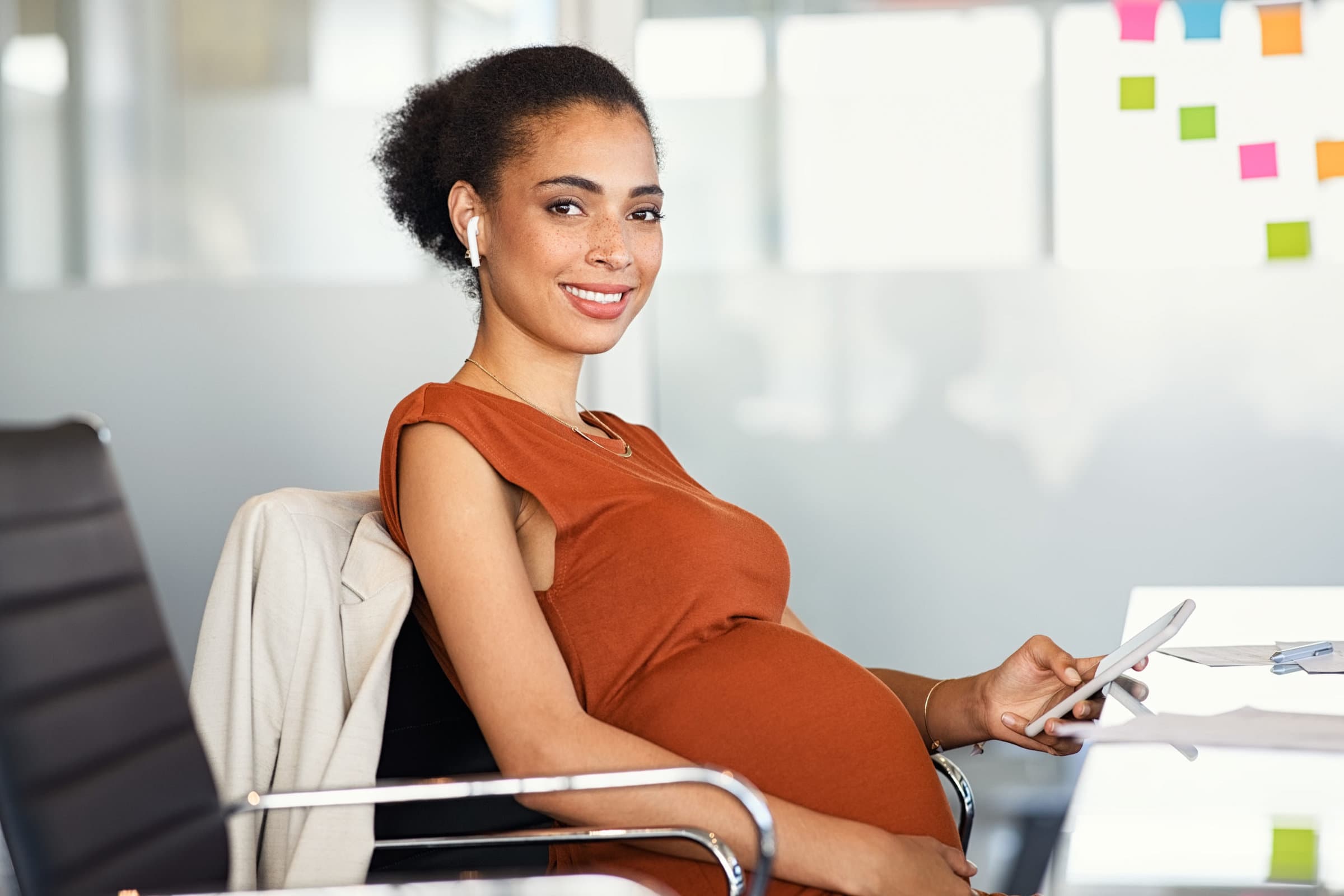 Portrait of smiling pregnant business woman using smartphone and doing video call with wireless earphone in modern office. Formal pregnant black businesswoman texting on phone in office while looking at camera. Portrait of happy african american mom expecting baby sitting in meeting room.