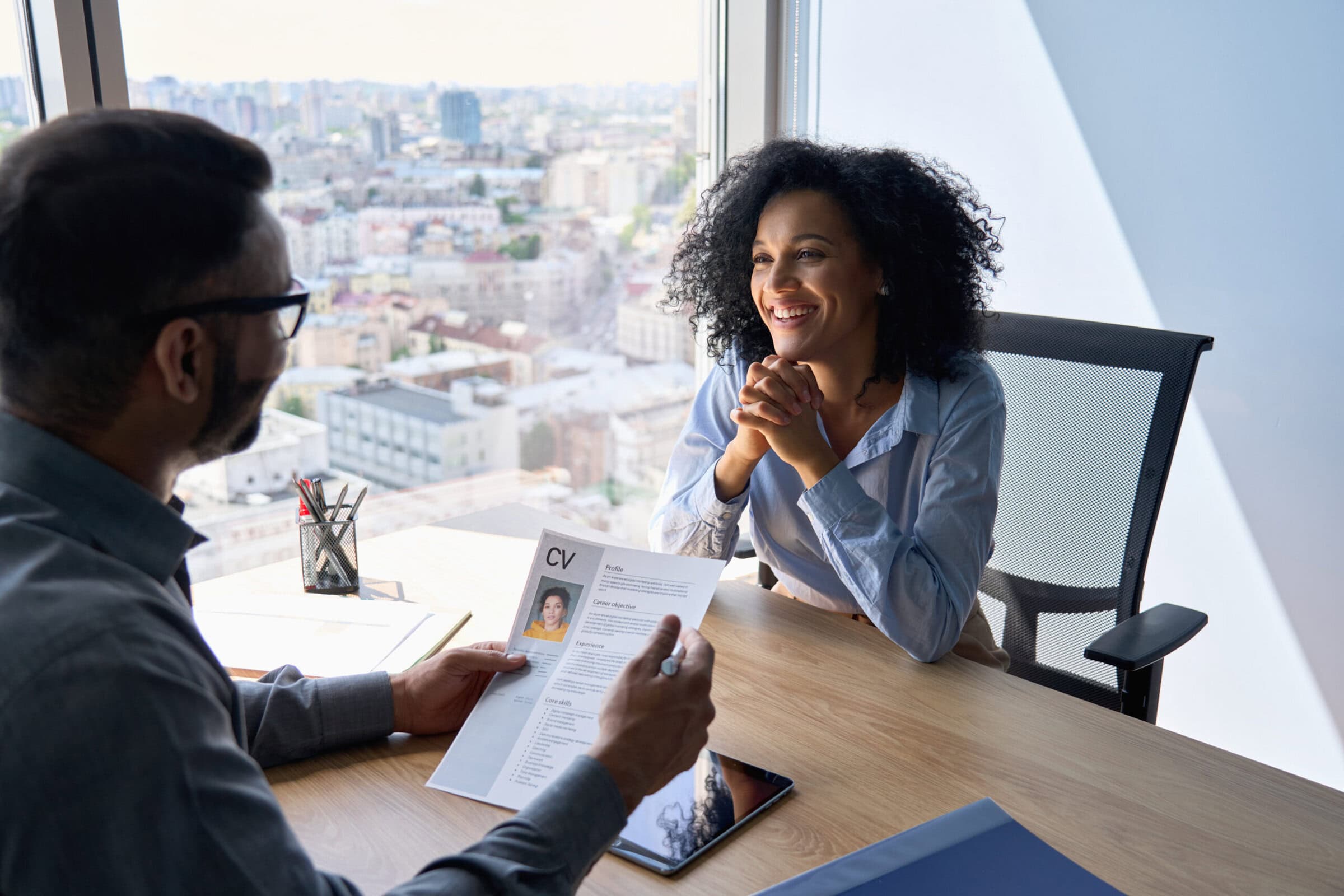 Friendly interview between indian hr manager and African American applicant. Friendly interview between Indian businessman hr director holding paper cv hiring for job female African American applicant manager sitting in contemporary office. Human resources recruitment concept.