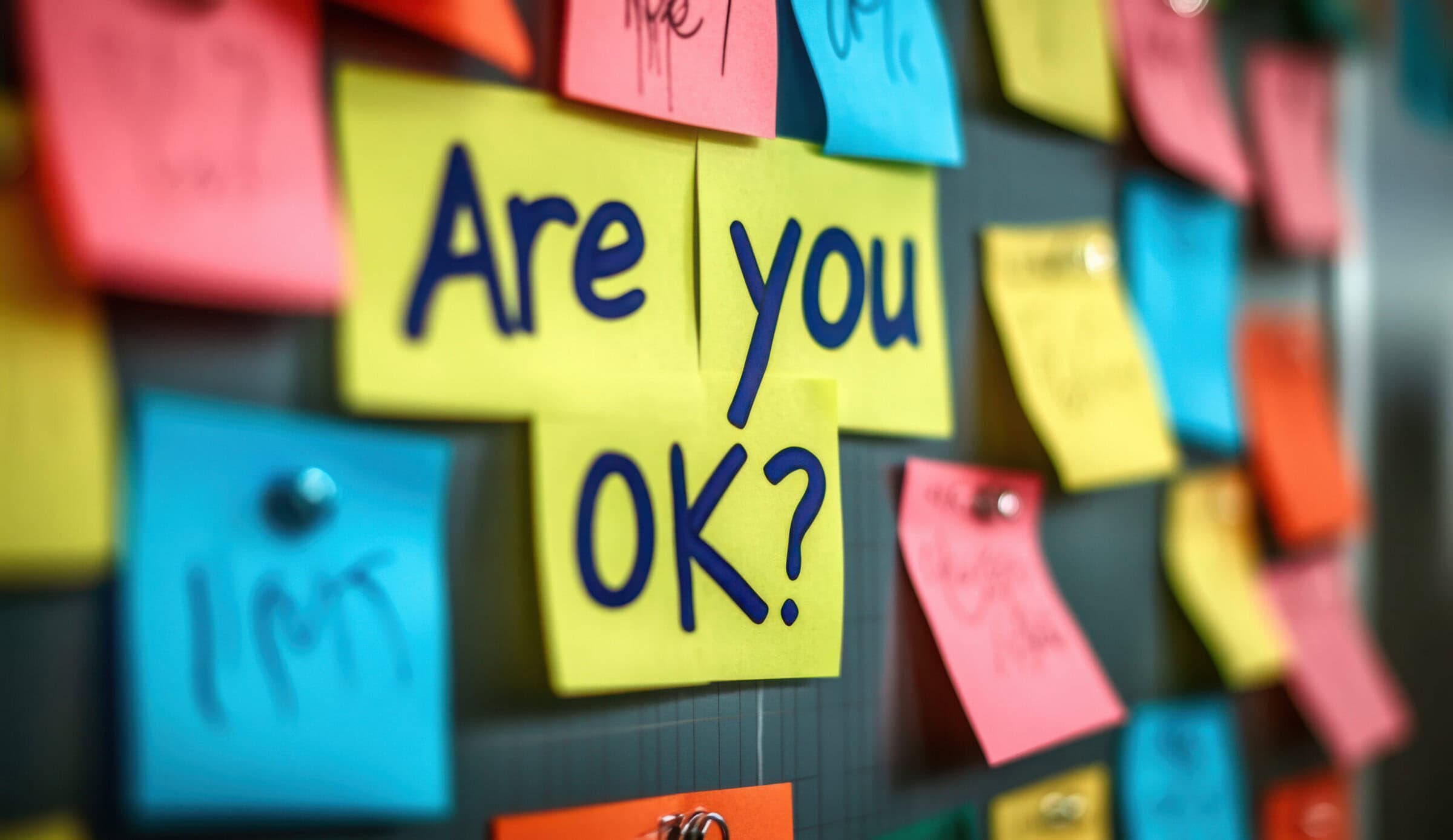 Colorful sticky notes on corkboard. One note says Okay Workplace mental health initiative. Employee support. Caring, encouraging work environment. Positive message for colleagues. Promoting Colorful sticky notes on corkboard. One note says Okay Workplace mental health initiative. Employee support. Caring, encouraging work environment. Positive message for colleagues. Promoting